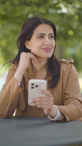 Vídeo-Vertical-De-Una-Mujer-Sentada-En-Una-Mesa-Al-Aire-Libre-En-Una-Calle-De-La-Ciudad-Mirando-El-Teléfono-Móvil